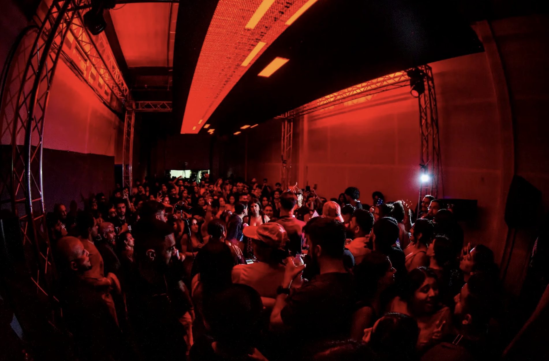 A packed dance floor under red light, trusses framing the crowd