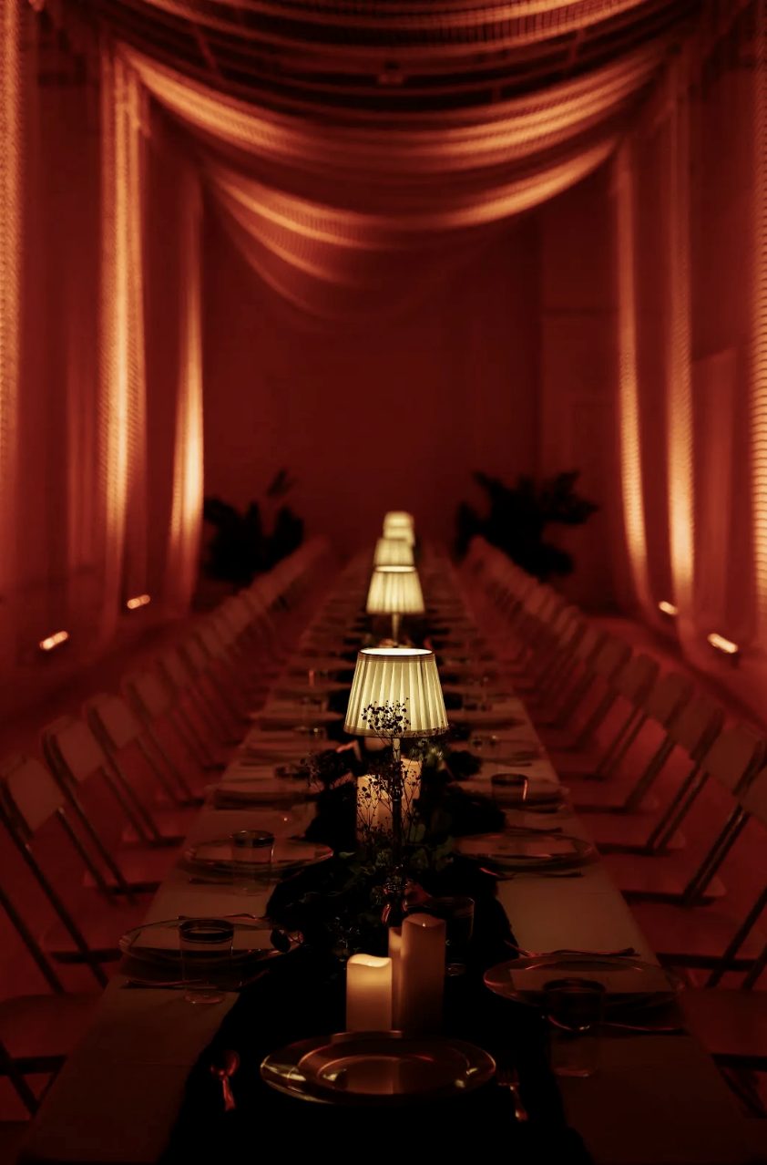 A lamplit long table set for a private dinner, the room washed in deep red